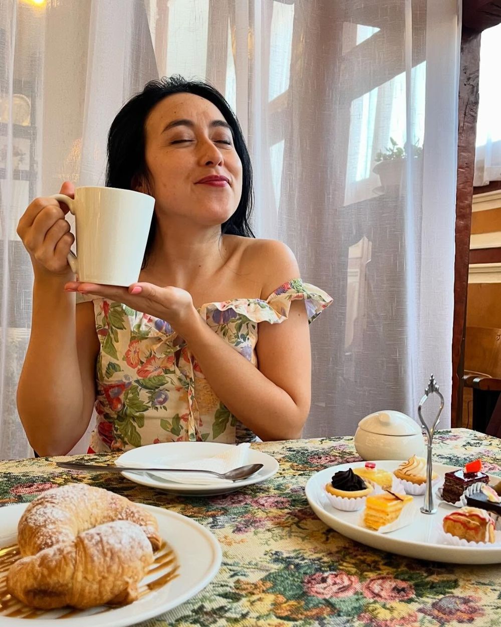 Mesa servida con té, croissant y pastelería sobre mantel floreado.